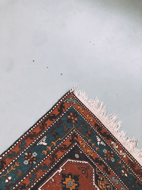 Close-up view of a corner of a traditional patterned rug with red, blue, and cream geometric designs and fringed edges, placed on a smooth, light grey floor. The surface appears clean and well-maintained, with the intricate details of the rug clearly visible. Visible in the background is an adjacent plain grey area, indicating a domestic or commercial space. The lighting is even, highlighting the vibrant colors and neat condition of the rug, which has likely been cleaned or properly maintained by Carpet Cleaning Harringay as part of their deep cleaning and sanitisation services for domestic spaces, specifically targeting rug and surface cleaning on Green Lanes, Harringay.