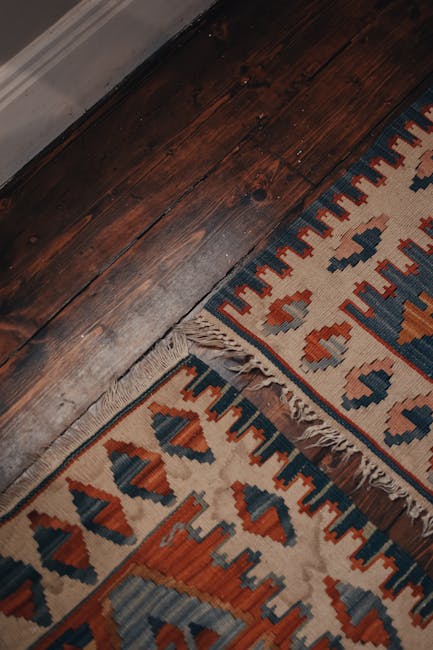 Close-up view of a corner of a traditional patterned rug with red, blue, and cream geometric designs and fringed edges, placed on a smooth, light grey floor. The surface appears clean and well-maintained, with the intricate details of the rug clearly visible. Visible in the background is an adjacent plain grey area, indicating a domestic or commercial space. The lighting is even, highlighting the vibrant colors and neat condition of the rug, which has likely been cleaned or properly maintained by Carpet Cleaning Harringay as part of their deep cleaning and sanitisation services for domestic spaces, specifically targeting rug and surface cleaning on Green Lanes, Harringay.