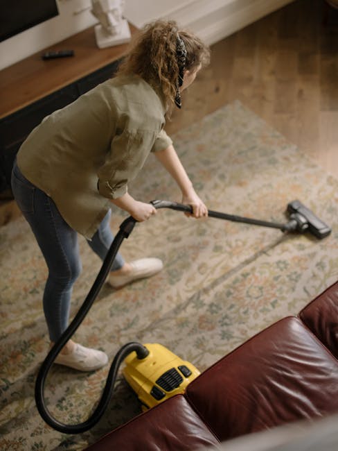 A woman with curly hair wearing a beige jacket, jeans, and white sneakers uses a yellow and black vacuum cleaner with a long hose attachment to deep clean and sanitise a patterned area rug in a living room. The room features a wooden floor, a dark wood sideboard with decorative items, and natural lighting. She concentrates on surface cleaning the rug, which appears free of dust and stains, contributing to the overall hygiene and cleanliness of the space. This image illustrates professional domestic cleaning services offered by Carpet Cleaning Harringay, highlighting their expertise in deep cleaning and maintaining pristine home environments.