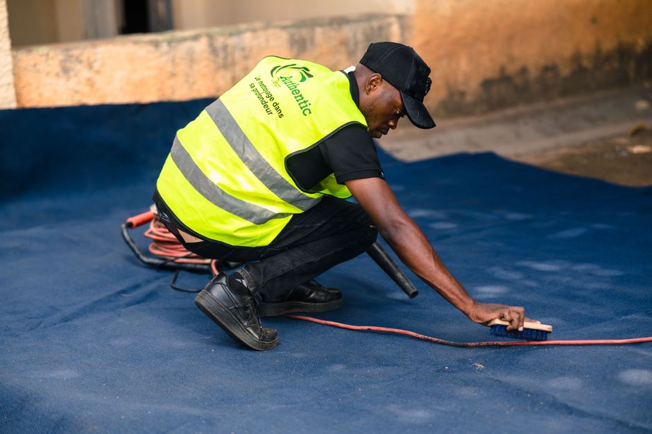 A professional cleaner wearing a high-visibility yellow vest with the logo and text 'Authentic' on the back, black cap, black t-shirt, and dark jeans is kneeling on a blue carpeted floor in an indoor 