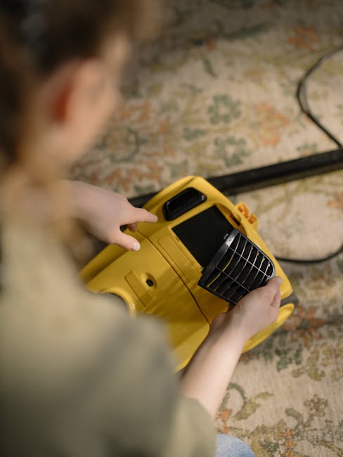 A professional cleaner wearing a high-visibility yellow vest with the logo and text 'Authentic' on the back, black cap, black t-shirt, and dark jeans is kneeling on a blue carpeted floor in an indoor 