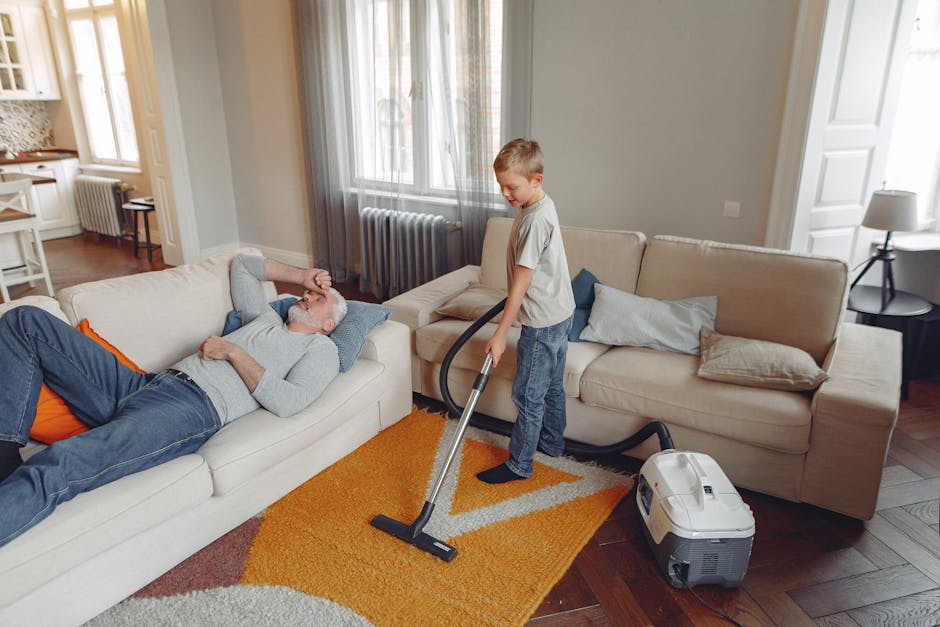 A person kneeling on a patterned carpeted floor, using a yellow and black portable vacuum cleaner for surface cleaning. The individual is dressed in casual clothing, including a beige jacket and blue 