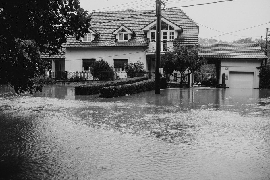 A two-story residential house on Wightman Road in Harringay, partially submerged in floodwater caused by heavy rain. The house has a tiled pitched roof with dormer windows, a small front garden with shrubs, and a driveway leading to a garage. Floodwaters cover the front yard and extend onto the street, reflecting the house and trees. Nearby trees and utility poles are visible, and the scene appears overcast, emphasizing the extent of the flooding. This image illustrates the impact of heavy flooding in the Harringay area, relevant to emergency flood response and cleaning services by Carpet Cleaning Harringay.
