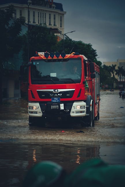 A flooded street near Wightman Road in Harringay during a heavy rainstorm, with water covering the asphalt pavement and partially submerging parked cars, including a silver vehicle in the center. Surrounding trees with green and yellow leaves are partially submerged, and the scene appears overcast with diffused natural light, highlighting the extent of flooding. The image reflects a recent flood event, emphasizing the importance of surface and deep cleaning for property restoration. The area is outdoors with visible water accumulation on the road surface, and no indoor surfaces or cleaning tools are present. The scene underscores the need for professional flood recovery and sanitisation services, like those offered by Carpet Cleaning Harringay.