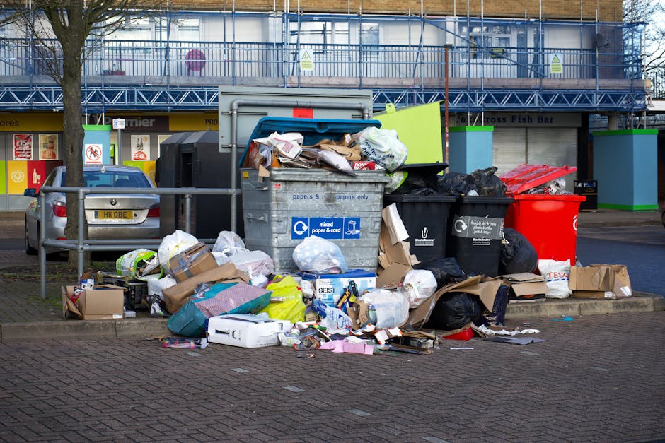 A collection of overflowing rubbish bins and scattered waste such as cardboard boxes, plastic bags, paper, and packaging materials, situated on a paved sidewalk in front of a commercial area with storefronts and a blue metal fence. The waste appears disorganized, with some trash spilling onto the ground, indicating a need for proper waste disposal or collection. The background includes a modern building with glass windows and metal framework, and a parked car nearby. The scene reflects typical urban waste accumulation outside retail outlets or public spaces, illustrating the importance of waste management and hygienic surface cleaning, as emphasized by Carpet Cleaning Harringay in relation to the rules for waste from upholstery cleaning, Haringey.