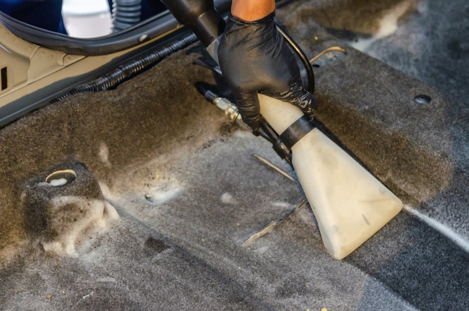 A person wearing black gloves operates a handheld upholstery cleaning device, applying deep cleaning and sanitisation to the grey carpeting of a vehicle interior. The device emits a spray of cleaning solution and rinses away dirt and stains, leaving the fabric damp and fresh. Multiple foam residues and wet patches are visible on the textured carpet surface, which shows signs of being in a vehicle, with nearby wiring and seat mounting points. Bright, even lighting highlights the cleanliness process taking place under the vehicle's seat area. This image demonstrates professional surface cleaning techniques used by Carpet Cleaning Harringay to maintain hygiene standards in both domestic and commercial vehicles, in line with Haringey Council rules for waste from upholstery cleaning.