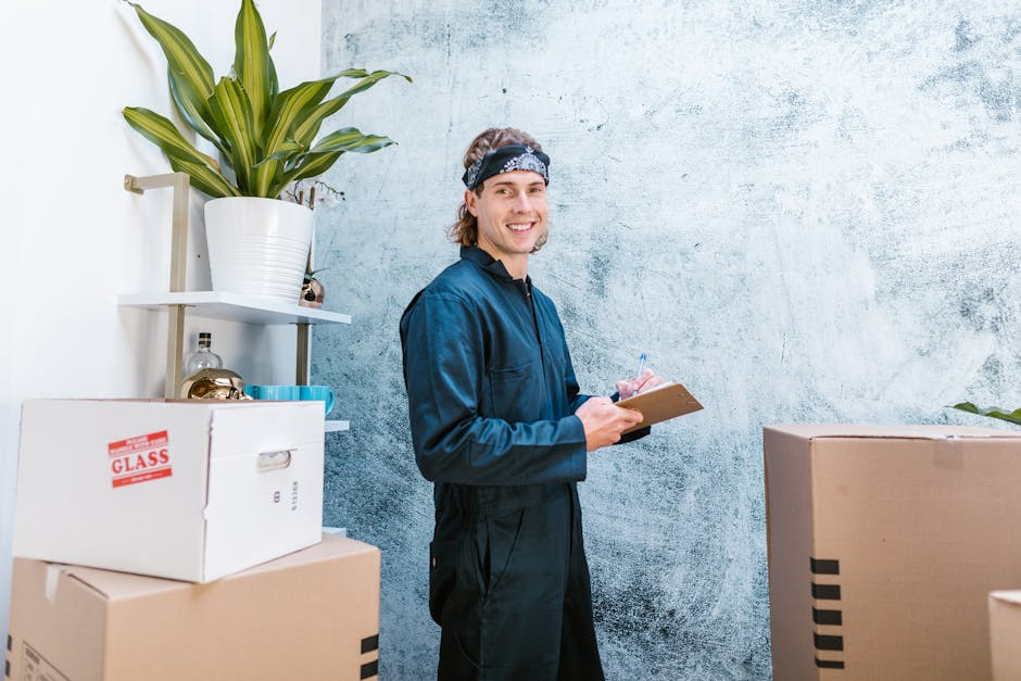 A young male cleaner with shoulder-length light brown hair, wearing a dark blue uniform and a patterned headband, stands in a room with a textured light blue wall. He is smiling and holding a clipboard with a sheet of paper and a pen, appearing to be inspecting or taking notes during a professional cleaning service. To his left, there are cardboard boxes stacked, one labeled 'GLASS,' and behind him, a white shelf holds a potted plant with large green leaves and a small decorative object. The scene is well-lit with natural or soft light, indicating a clean and organized environment suitable for surface cleaning or deep cleaning in a residential or commercial space, as provided by Carpet Cleaning Harringay.