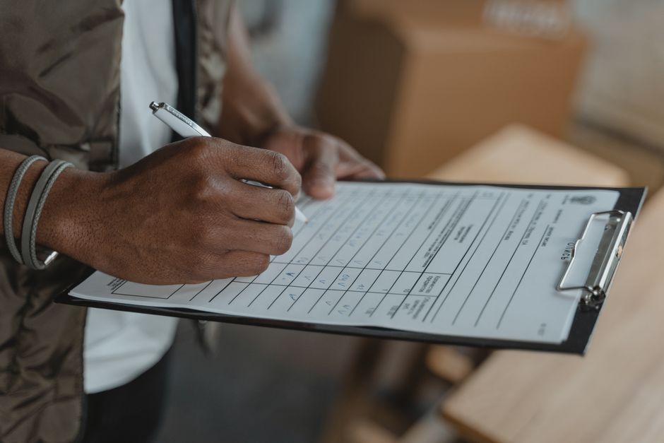 Close-up of a person's hand holding a pen and writing on a form attached to a black clipboard. The individual is wearing a grey wristband and a brown jacket, with part of their arm visible. The background includes blurred elements of a room with a cardboard box and possibly furniture or fixtures, indicating an indoor setting. The lighting appears natural, illuminating the workspace evenly. This image relates to the detailed process of conducting an end-of-tenancy check, which is essential for thorough internal cleaning and sanitisation services, as offered by Carpet Cleaning Harringay in accordance with landlord checklists and rules for property maintenance.