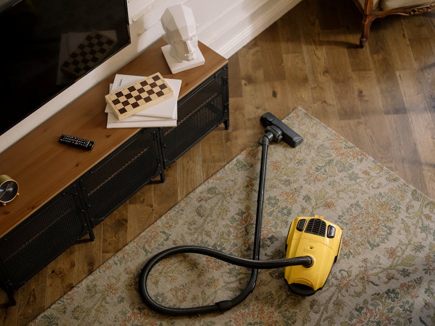 A woman with curly hair, dressed in a beige jacket, dark jeans, and white shoes, is engaged in surface cleaning by vacuuming a patterned area rug in a living room. The room features a wooden floor adjacent to the rug, and is illuminated by soft natural or artificial light. She holds a vacuum cleaner with a yellow body and a black hose, with the nozzle positioned on the rug, demonstrating a deep cleaning process. The background includes a wooden sideboard with decorative items, and part of a red leather sofa is visible in the foreground. This image illustrates domestic cleaning services provided by Carpet Cleaning Harringay, emphasizing thorough hygienic maintenance of home surfaces.