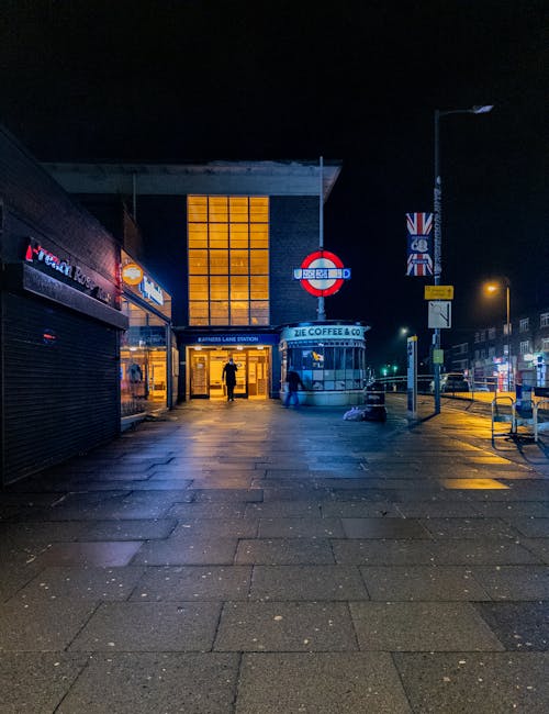 Nighttime view of the entrance to Turnpike Lane station in Harringay, featuring illuminated signage, a glass facade with warm lighting, and a paved footpath with wet, reflective surfaces. A person is visible walking towards the station entrance, with streetlights casting a yellow glow on the surrounding area. Adjacent shops are closed, their shutters down, and a lamppost stands on the right side of the image. This scene showcases urban cleanliness and safe pedestrian access near the station, as captured in a clear, well-lit photograph. Carpet Cleaning Harringay offers surface cleaning and deep cleaning services to maintain hygiene and cleanliness in residential and commercial spaces.