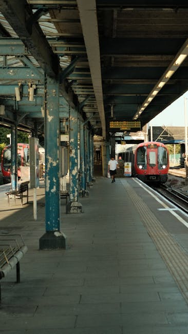 The image depicts a train station platform with a metal canopy supported by weathered blue-painted columns. The platform surface is paved with grey tiles, appearing clean and well-maintained, with tactile paving for visually impaired passengers running parallel to the train tracks. A person is seen walking away from the camera towards a red train that is stationary on the right side of the image. The station's lighting is provided by mounted overhead lights, and the overall environment appears orderly and hygienic. Carpet Cleaning Harringay specializes in surface cleaning and deep cleaning services, ensuring hygienic and sanitized environments for both residential and commercial spaces, similar to maintaining cleanliness in public transport stations.
